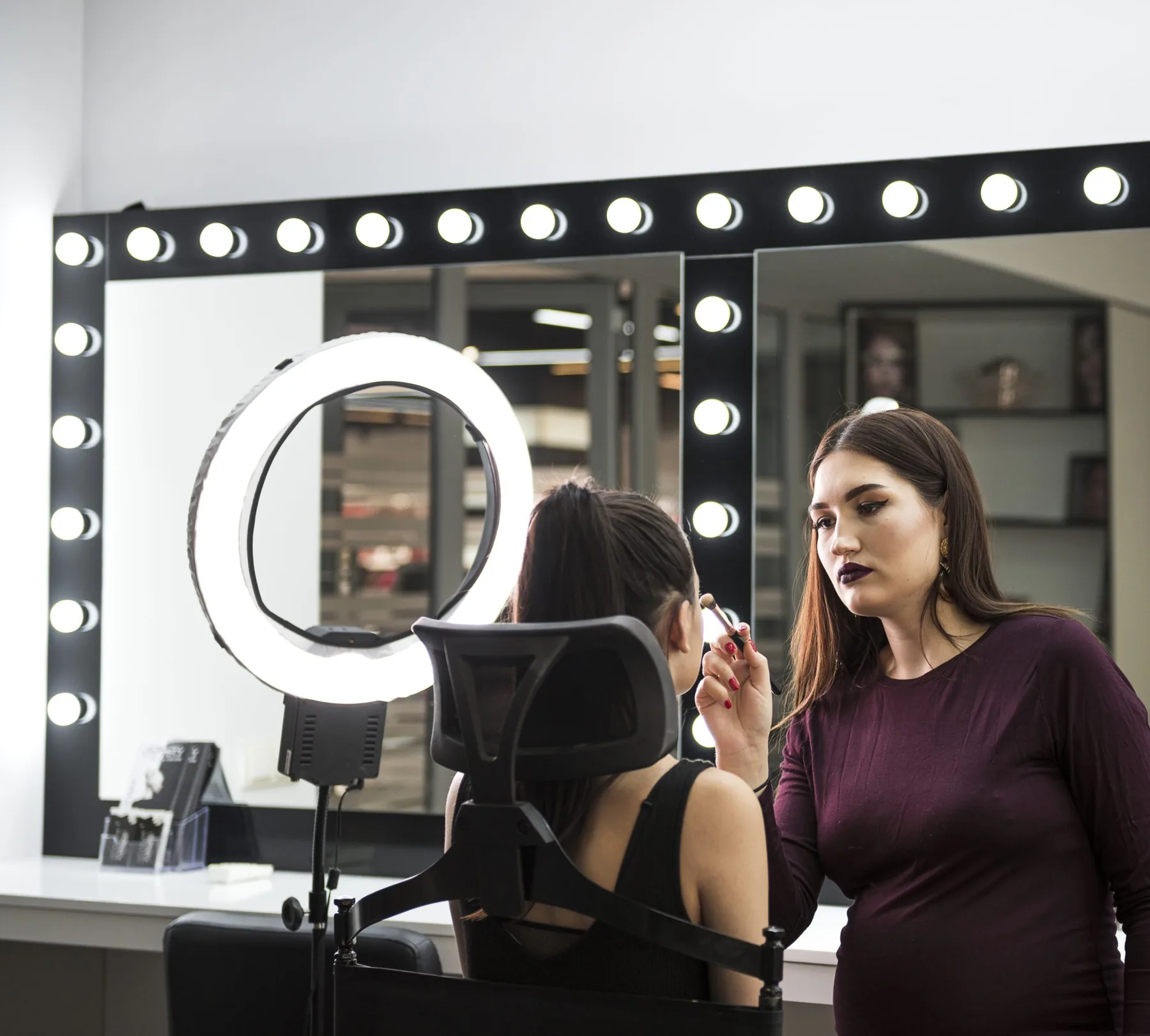 A make-up artist applying make-up to a client at a lit salon mirror